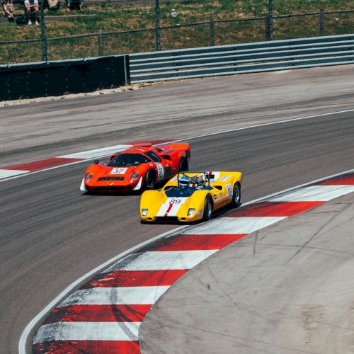 On a racetrack, a bright yellow race car leads an orange and red sports car through a turn, close together.