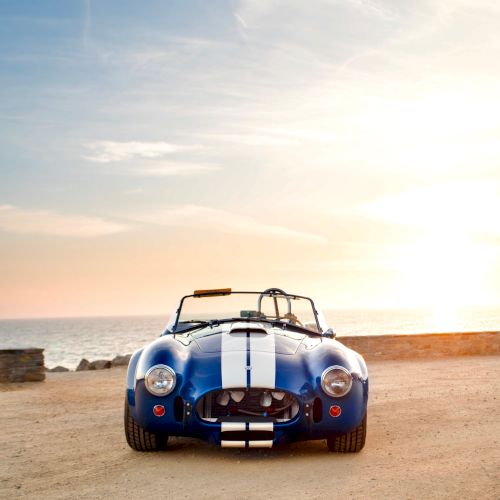 A blue vintage sports car sits on a sandy beach at sunset with the ocean and rocks in the background.