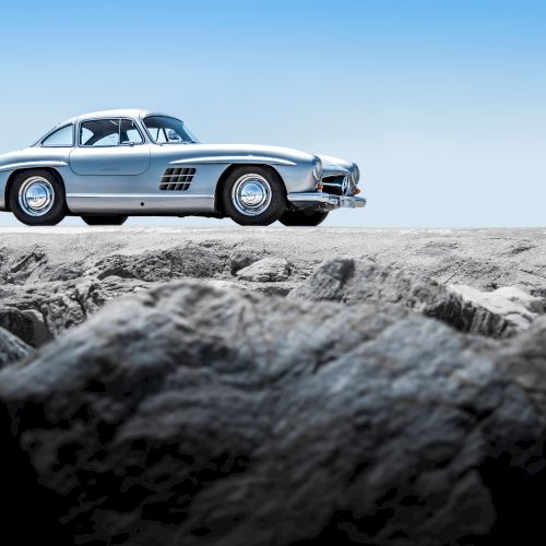 A classic silver Mercedes coupe drives across rocky terrain under a clear blue sky.