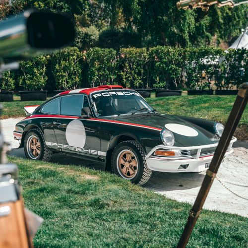 A classic Porsche coupe with red-and-green stripes sits on a sunny lawn by hedges and a fountain, gleaming outdoors.