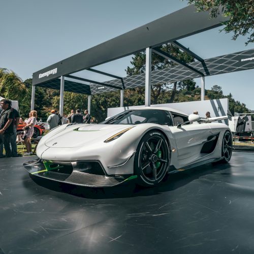 A sleek white-and-green supercar sits on display under a solar-paneled canopy at an outdoor event.