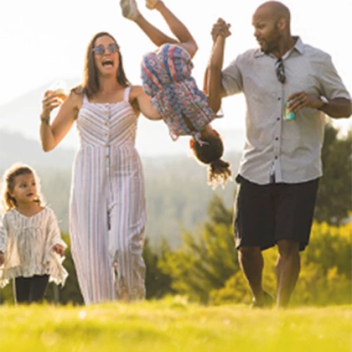 A family of four enjoys a sunny day outdoors; the parents swing one child in the air while the other walks alongside them.