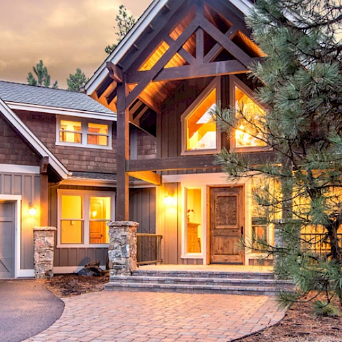 A modern, large house with a well-lit exterior, double garage, stone accents, and surrounded by trees at dusk.