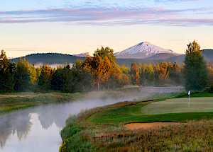 A scenic view of a golf course beside a river, with a snowy mountain and trees in the background under a partly cloudy sky.