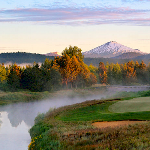A scenic view of a golf course beside a river, with a snowy mountain and trees in the background under a partly cloudy sky.