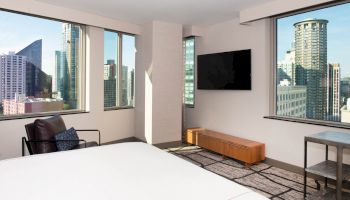 This image shows a modern hotel room with large windows offering a cityscape view, a TV on the wall, a chair, and a bed.
