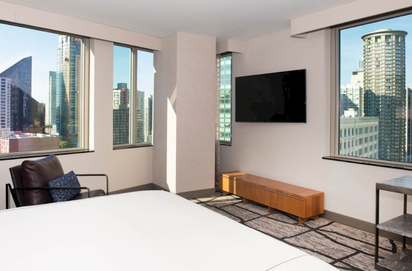 This image shows a modern hotel room with large windows offering a cityscape view, a TV on the wall, a chair, and a bed.