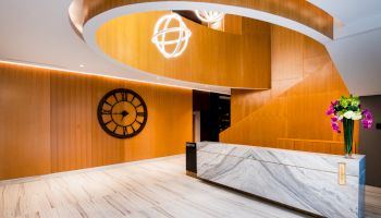 The image shows a sleek modern reception area with a spiral wooden structure, a large clock on the wall, and a marble desk with flowers.