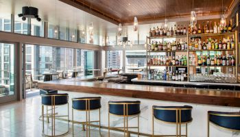 A sleek, modern bar area with a polished wooden counter, high stools, and a well-stocked shelf of various bottles. Large windows provide city views.