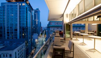 A modern cityscape with tall buildings at dusk, featuring a stylish balcony with tables, chairs, and a view of the street below.