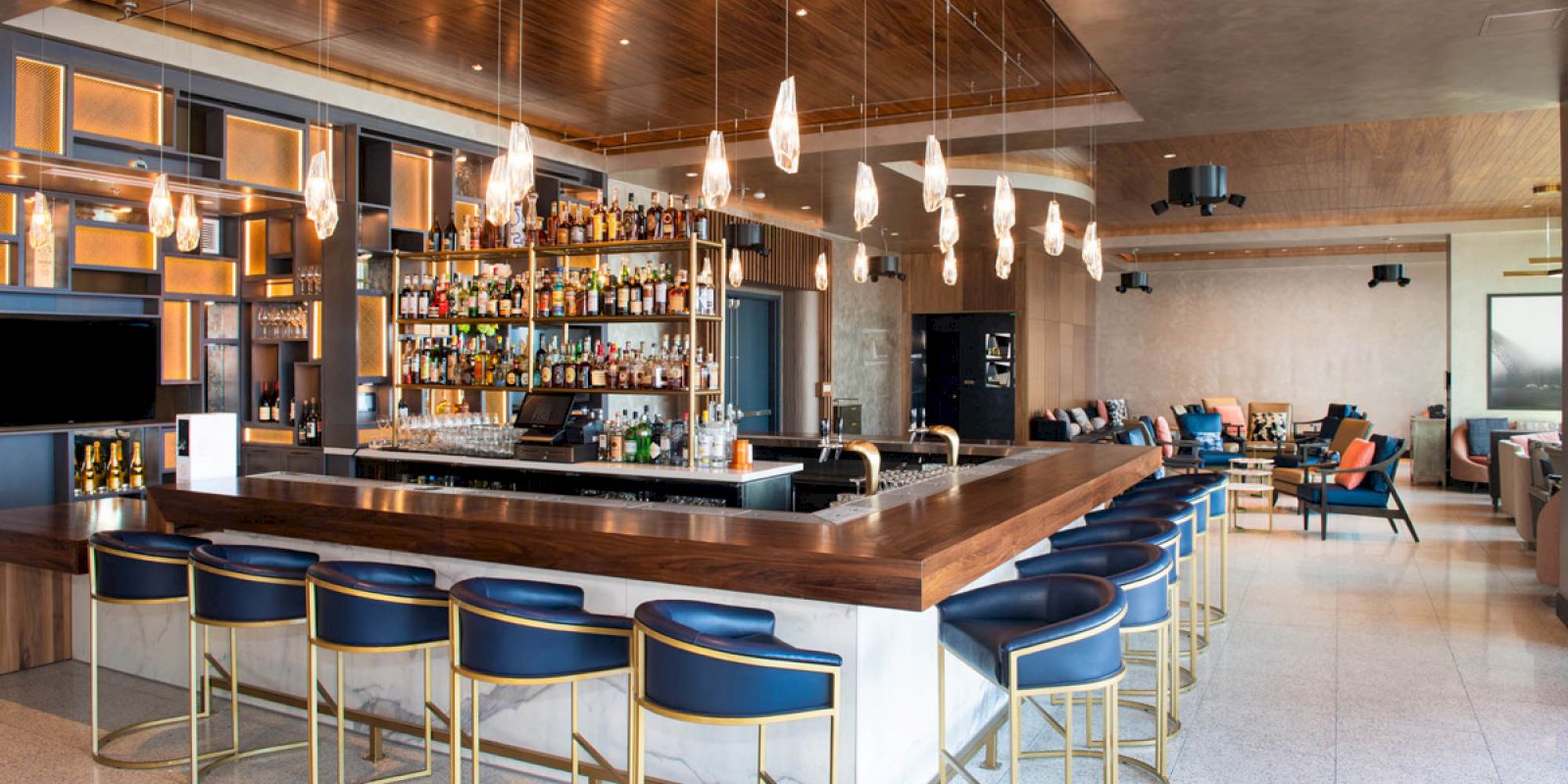 A modern bar with stylish lighting, a variety of liquor bottles, blue bar stools around a wooden counter, and seating areas in the background.