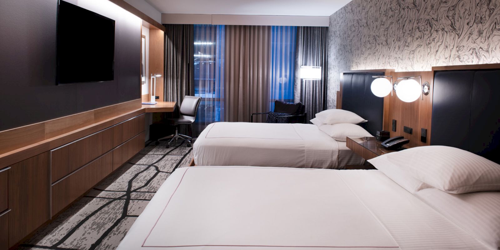 The image shows a modern hotel room with two beds, a flat-screen TV, a desk with a chair, and large windows with curtains.