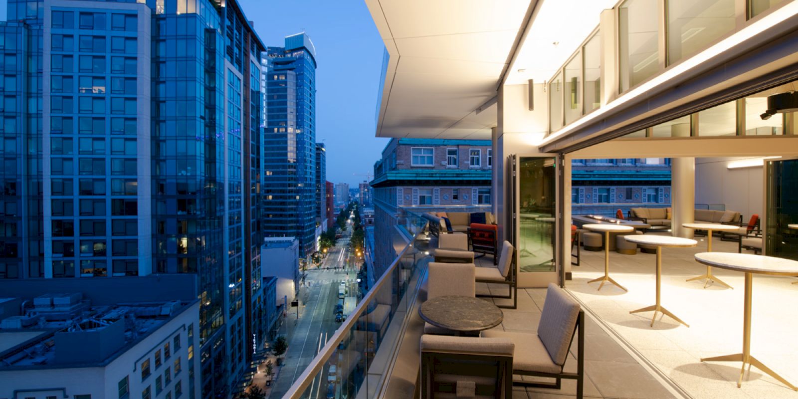 The image shows a modern rooftop terrace with outdoor seating, overlooking a cityscape of tall buildings at dusk.
