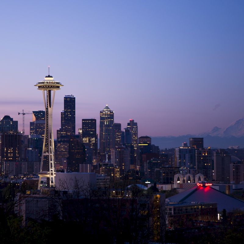 The image shows the Seattle skyline at dusk with the Space Needle prominently featured and Mount Rainier in the background, under a twilight sky.