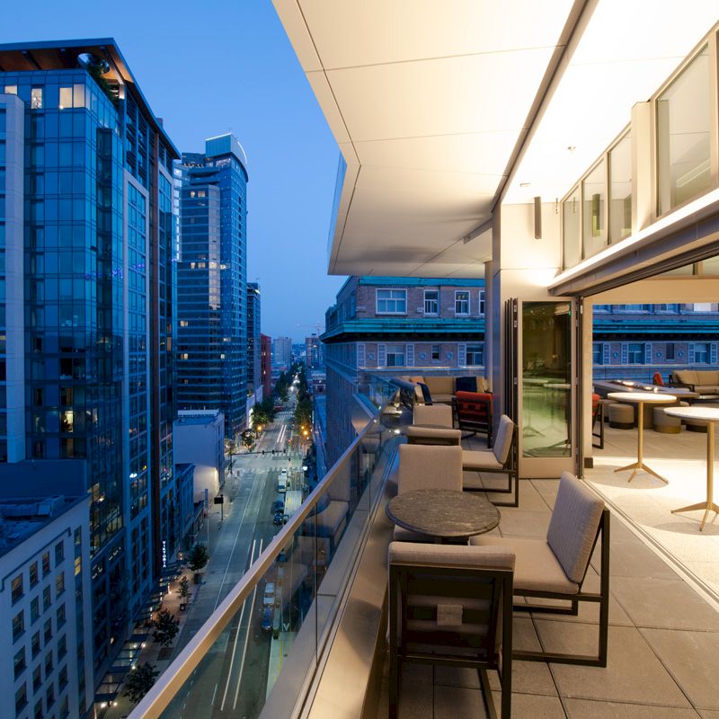 The image shows a modern city view from a high-rise balcony at dusk, with outdoor seating and a contemporary open indoor space.