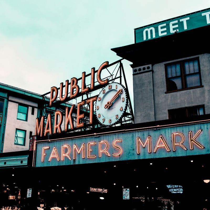 The image features the iconic neon sign of the Public Market Center and Farmers Market in an urban setting, with surrounding buildings and signage.