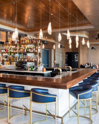 This image shows a modern bar with a wooden counter surrounded by blue stools, hanging pendant lights, and a well-stocked back bar.