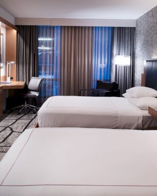 A modern hotel room with two neatly made beds, a wall-mounted TV, a desk with a chair, and stylish lighting fixtures.