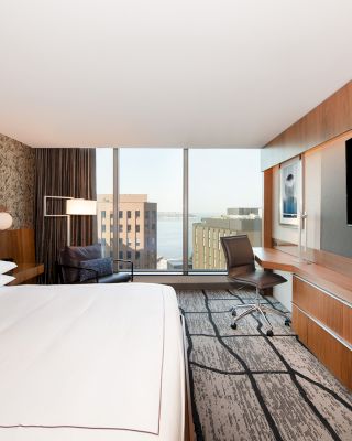 A modern hotel room featuring a large bed, a wall-mounted TV, a desk with a chair, a seating area, and a window with a city view.