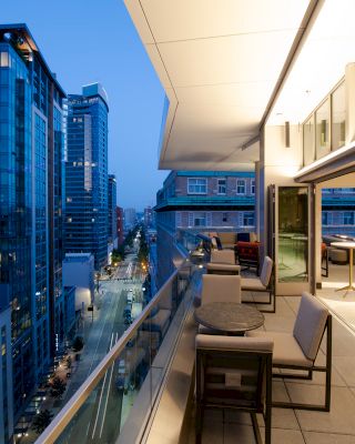 The image shows a modern urban balcony at dusk with tables and chairs, overlooking a city street lined with high-rise buildings.