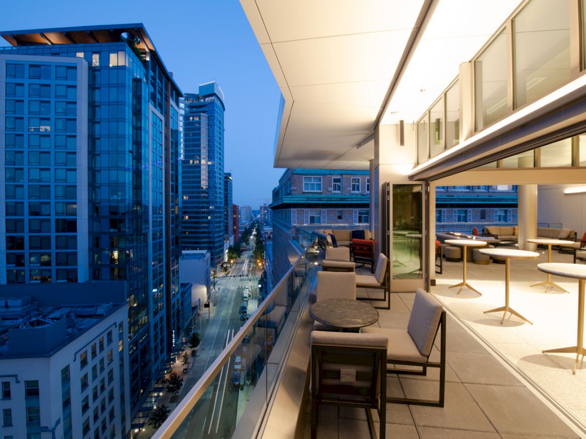 The image shows a modern cityscape at dusk with tall buildings and a stylish balcony with tables and chairs on the right side.