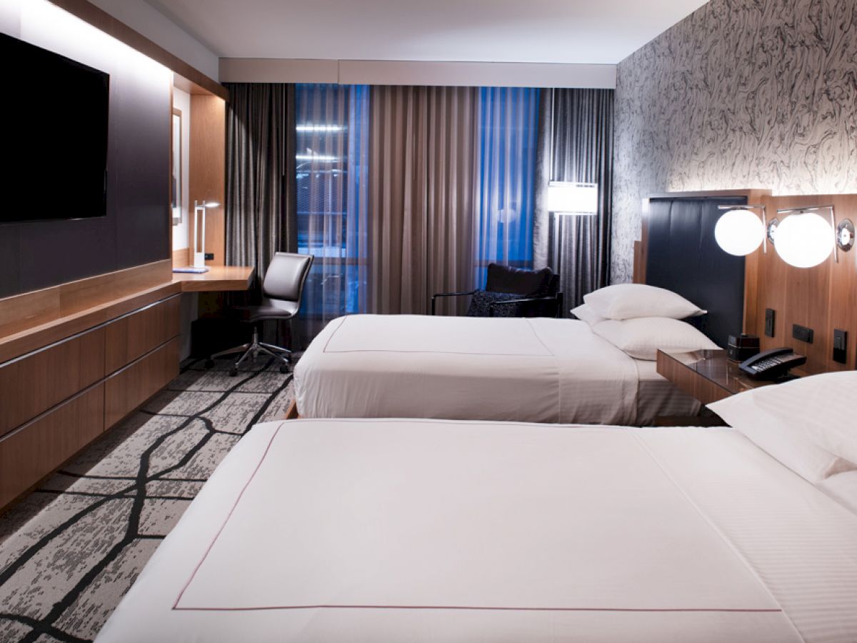 The image shows a modern hotel room with two beds, a TV, a desk with a chair, nightstands, and artistic wallpaper.