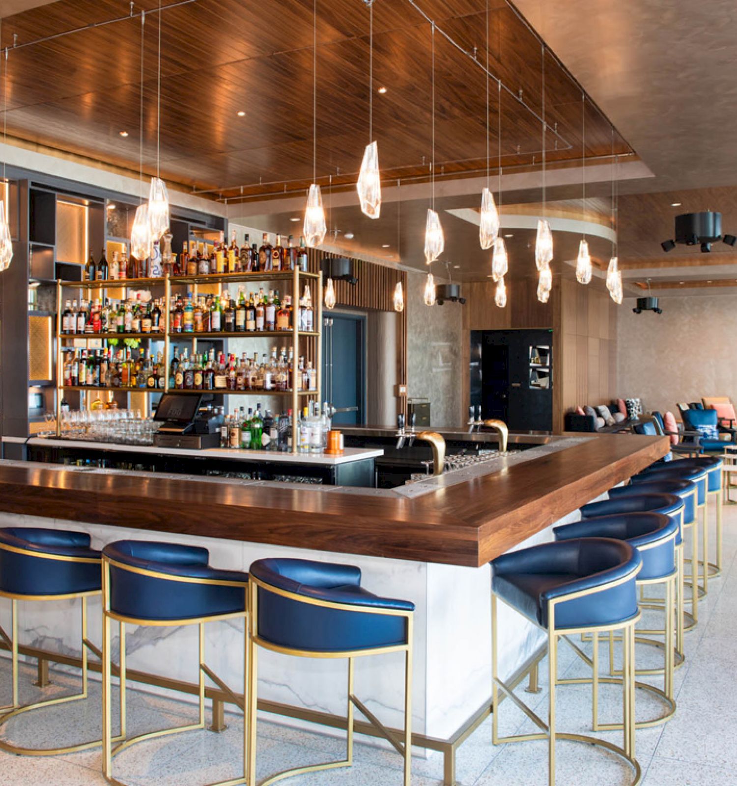 The image shows a modern bar with a long wooden counter, blue barstools, hanging lights, and shelves filled with bottles of liquor and glasses.
