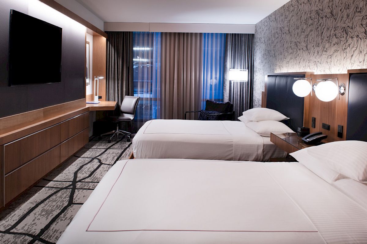 A modern hotel room features two neatly made beds, a wall-mounted TV, a study desk, and a patterned accent wall, creating a cozy and stylish atmosphere.