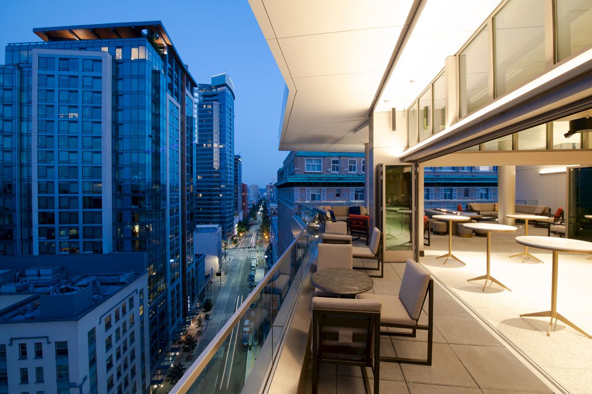 A modern balcony at dusk with tables, chairs, and an outdoor seating area overlooking a cityscape of tall buildings and a lit-up street below.