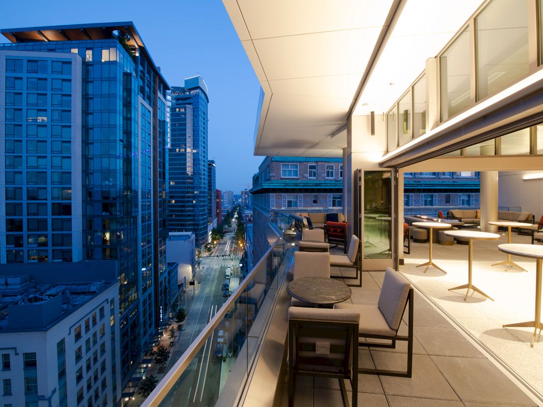 A modern balcony at dusk with tables, chairs, and an outdoor seating area overlooking a cityscape of tall buildings and a lit-up street below.