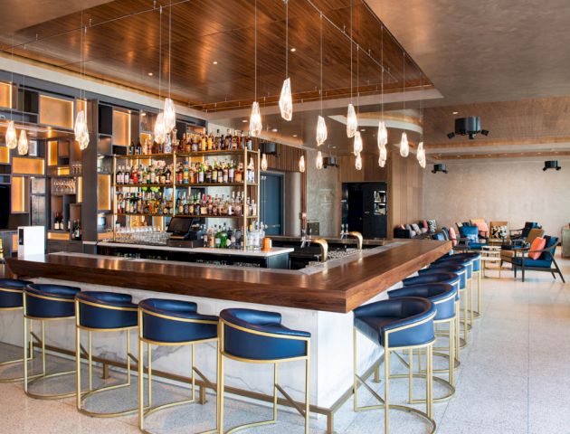 The image shows a modern bar with blue and gold bar stools, a fully stocked bar, hanging lights, and a seating area with chairs and a TV.