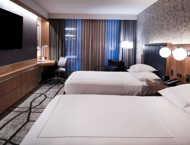 This image shows a modern hotel room with two beds, a desk and chair, a flat-screen TV, and a large window with curtains.
