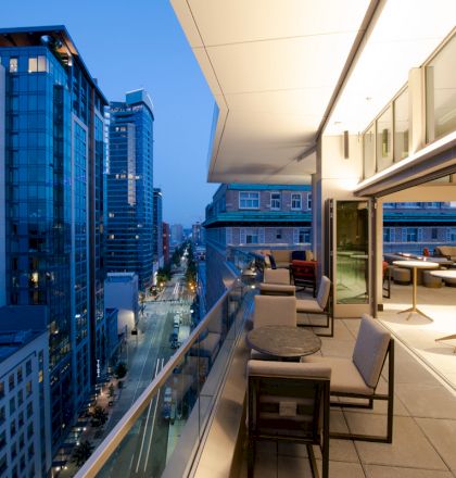 An outdoor terrace of a high-rise building overlooks a cityscape with modern buildings and streets, featuring seating and tables.