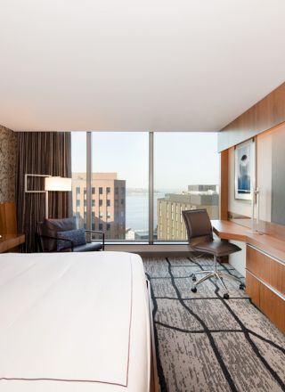 A modern hotel room with a large bed, TV, desk, chair, stylish decor, and a window overlooking a cityscape and a body of water ends the sentence.