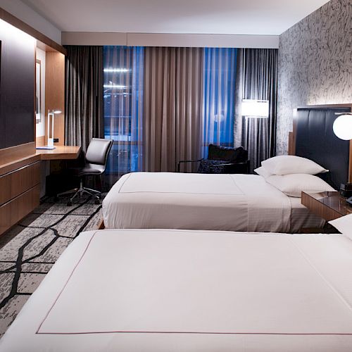 A modern hotel room with two beds, a TV, a desk with a chair, lamps, and patterned curtains on the window ends the sentence.
