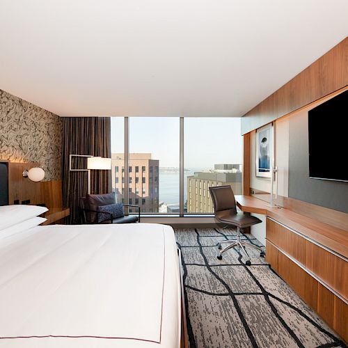 A modern hotel room with a large bed, a wall-mounted TV, a desk with a chair, and a window offering a city view.