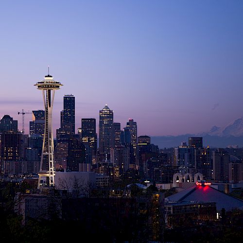A city skyline at dusk with prominent tall buildings, featuring the Space Needle and a distant mountain, likely Mount Rainier, in the background.