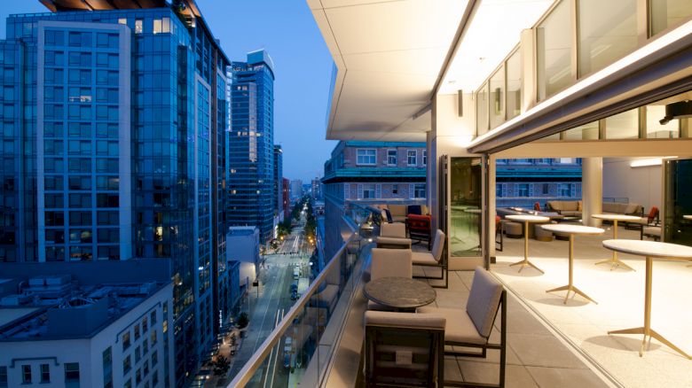 An elegant rooftop terrace at dusk overlooks a city street with modern high-rise buildings, featuring outdoor seating and well-lit tables.