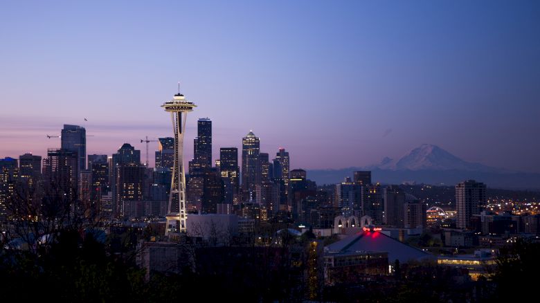 The image showcases the Seattle skyline at dusk, featuring the Space Needle prominently, with Mount Rainier visible in the background.