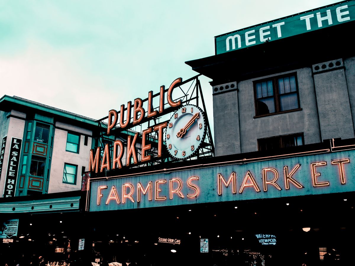 The image shows a neon-lit sign for a Public Market and Farmers Market at a well-known location with a clock above the sign and buildings in the background.
