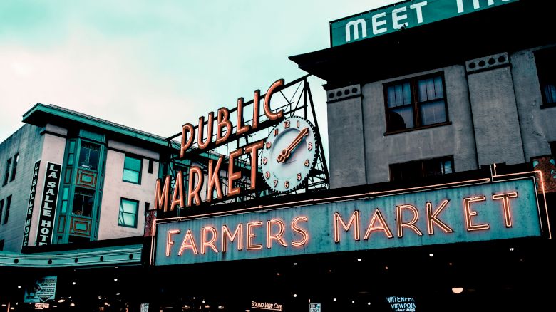 The image shows a neon-lit sign for a Public Market and Farmers Market at a well-known location with a clock above the sign and buildings in the background.