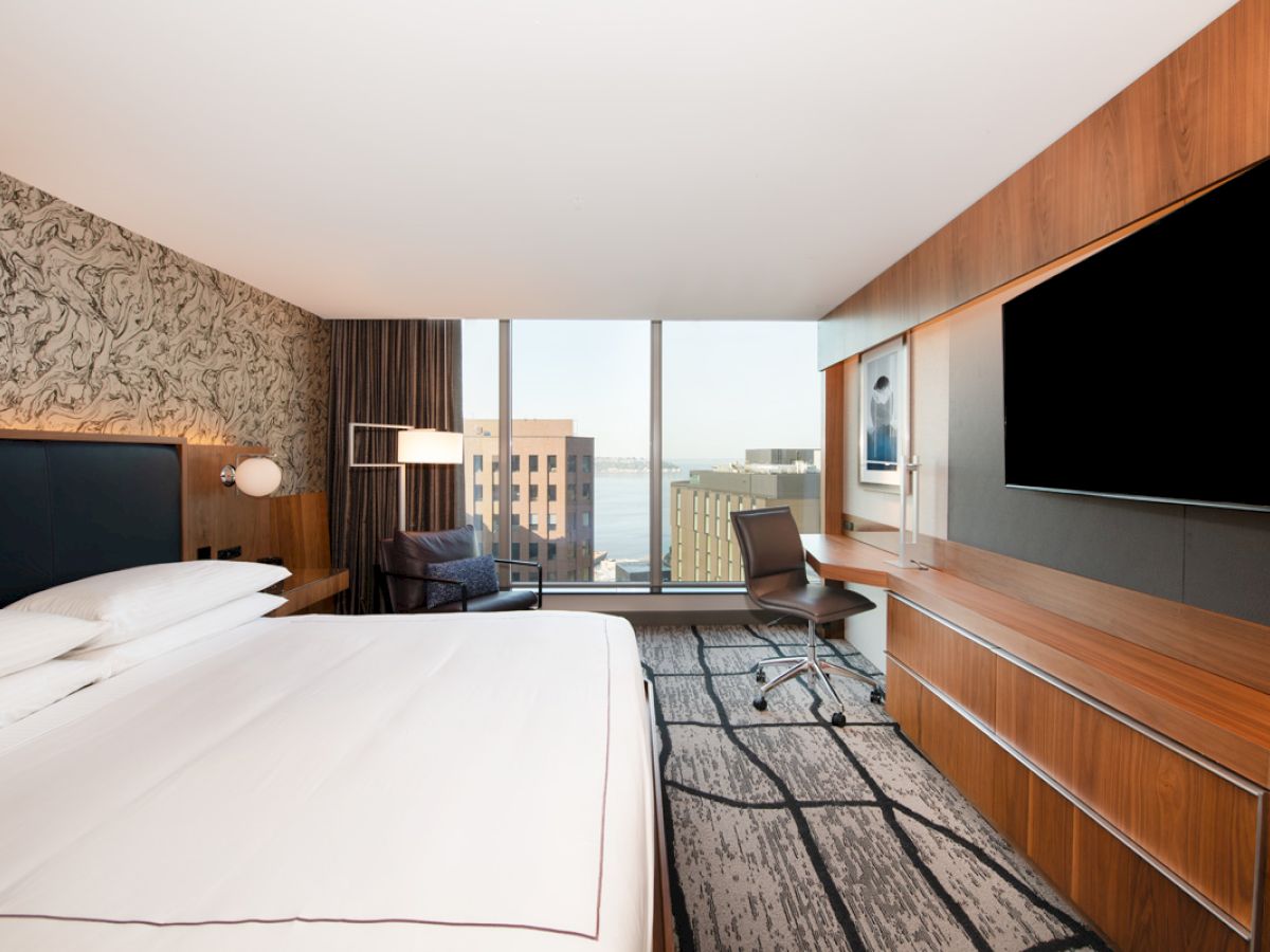 A modern hotel room with a large bed, wall-mounted TV, desk, chair, and a window view of buildings.