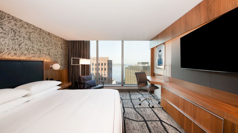 A modern hotel room with a large bed, wall-mounted TV, desk, chair, and a window view of buildings.