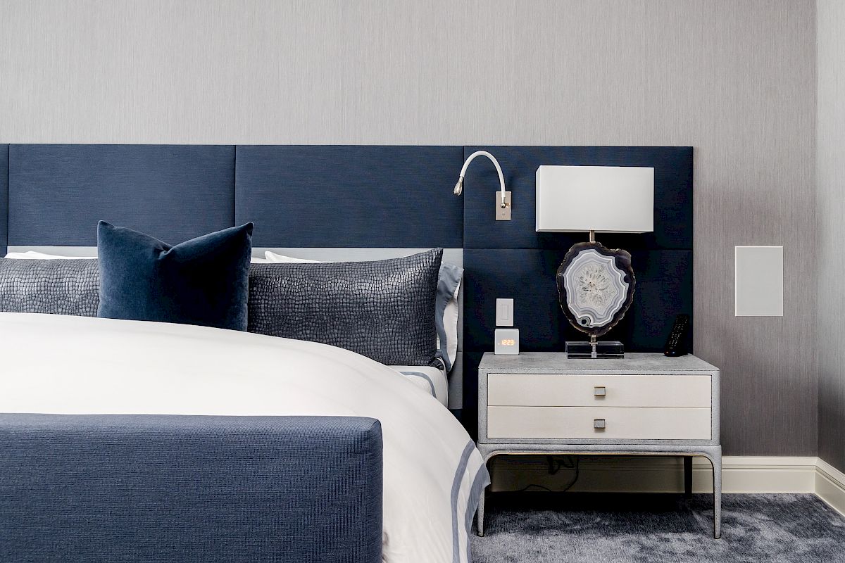 A modern bedroom with a blue upholstered headboard, white bed linens, a nightstand with a lamp, and a decorative item.