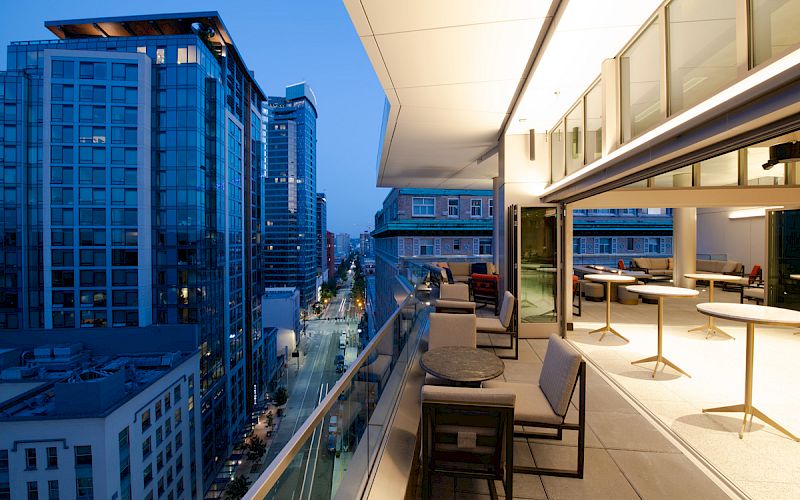 A modern rooftop terrace with seating overlooks a city street lined with tall buildings, captured at twilight just as the city lights begin to glow.