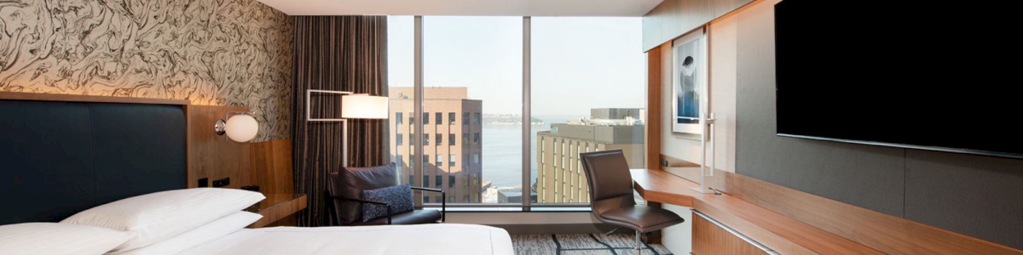 A modern hotel room with a bed, desk, chair, large TV, and a view of buildings from a window. The decor is sleek with wood accents.