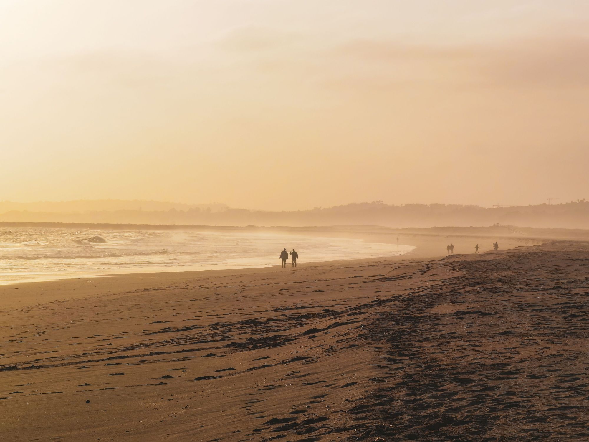 A serene beach at sunset with golden light, gentle waves, and a few distant walkers along the shoreline.